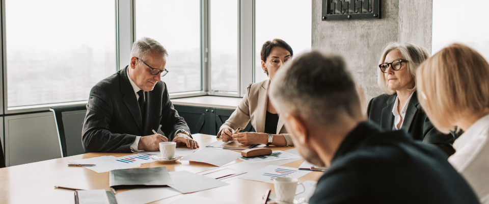 Five business professionals in a meeting around a conference table, reviewing printed documents and charts.